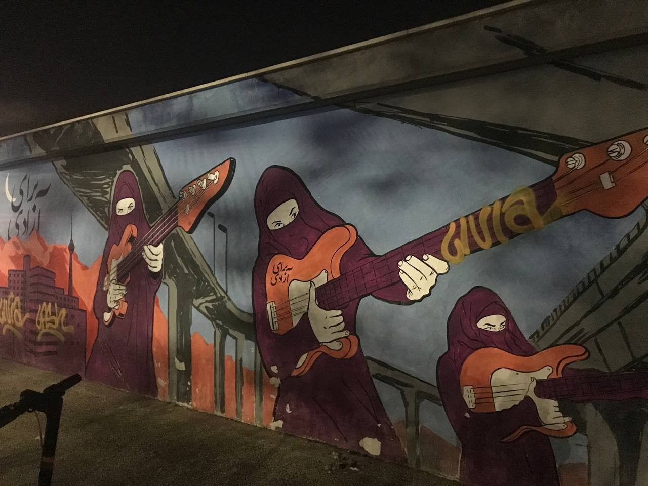 Street Art depicting 3 women in black blog attire rocking out with guitars. On the guitar of the woman in the middle and on the top left corner of the art is written "For Freedom" in Persian.