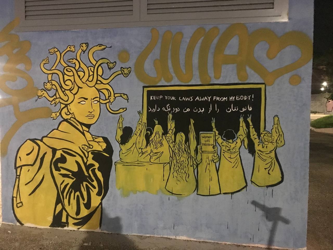 Street Art Depicts a schoolgirl with medusa snake hairs on the left of the frame. On the right is the recreation of a famous schoolgirls picture in a classroom without hijab, holding their hands out with peace signs. On the Classroom blackboard is written <br />"Keep your laws away from my Body" in English & Persian.