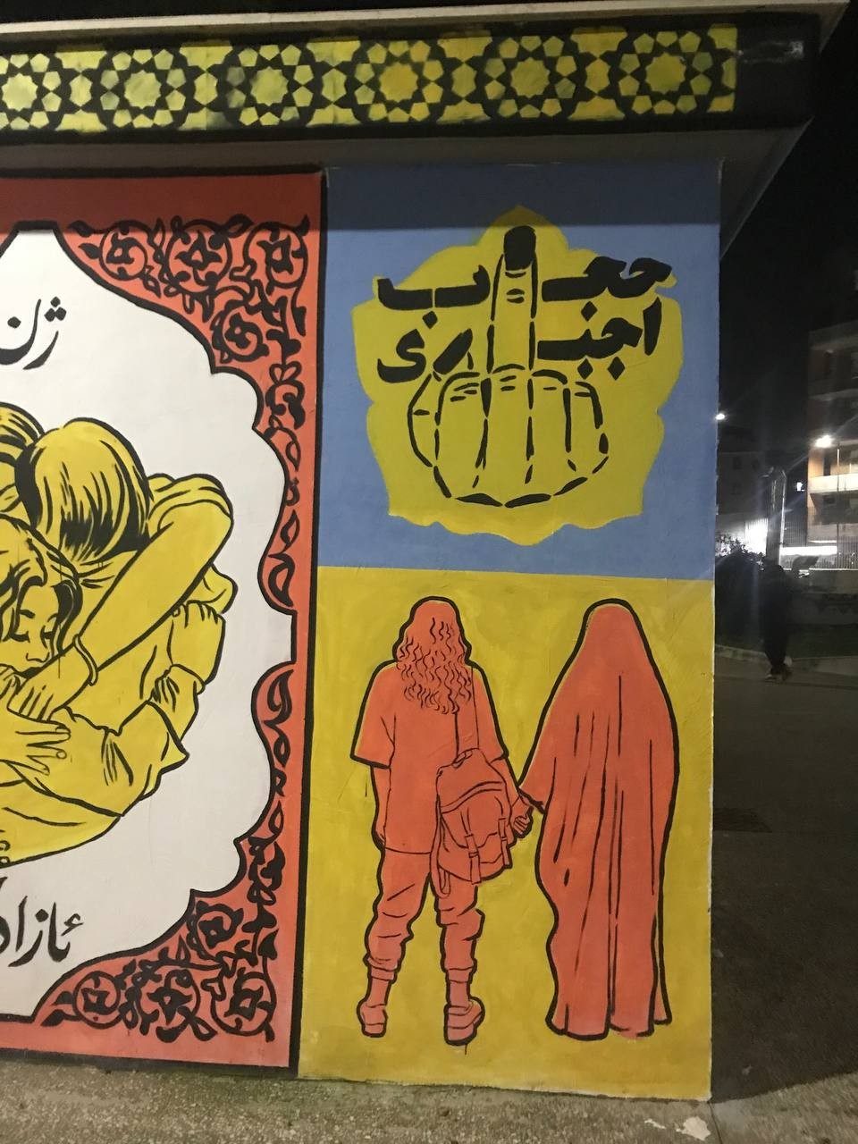 Street Art Depicting a middle finger to "Compulsory Hijab" Below it, two women, one with hijab and other without it, are holding hands in solidarity. On the left, there is picture of women hugging in solidarity, with "Woman Life Freedom" in Kurdish around <br />it.