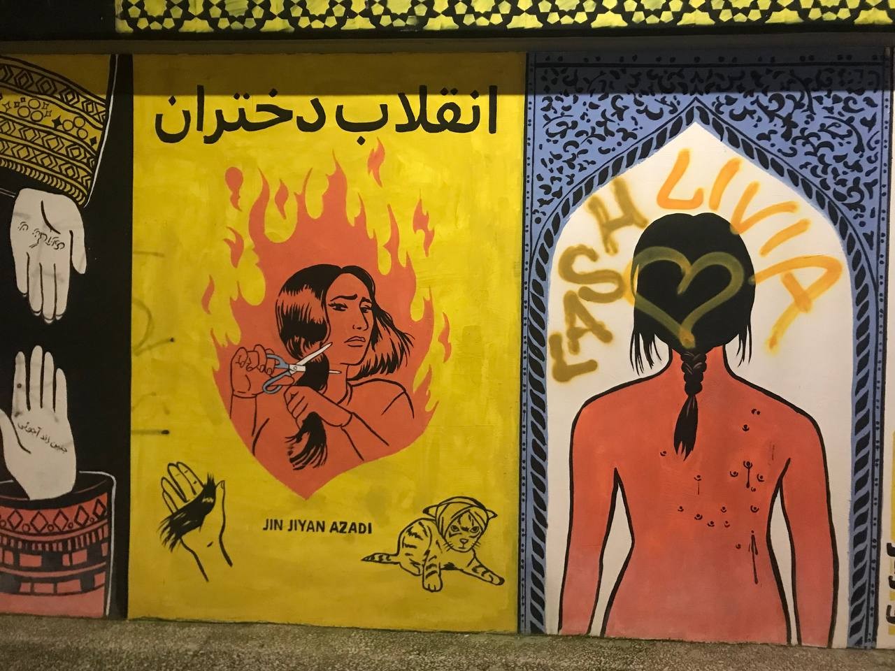 Street art depicts two hands, with one having "Woman Life Freedom" in Persian and the other in Baluchi. In the Middle, it depicts a woman cutting her hair in the fire of the revolt, and "Girls Revolution" on top is written in Persian, and "Jin Jiyan <br />Azadi" in Kurdish below it. On the right depicts a woman's back with anti-riot pallet wounds.